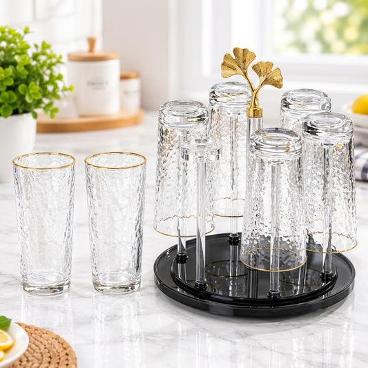 A set of clear glass drinking glasses with a hammered texture and gold trim, including two standing glasses and a six-piece inverted glass holder on a black round base with a gold leaf-shaped accent, displayed on a marble countertop in a bright kitchen setting.